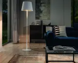 Biała, minimalistyczna lampa stojąca oświetla przytulny salon z granatową kanapą i szklanym stolikiem.